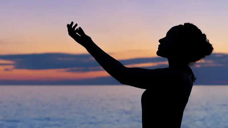 Silhouette of a person reaching gently toward a calm sunset sky over water